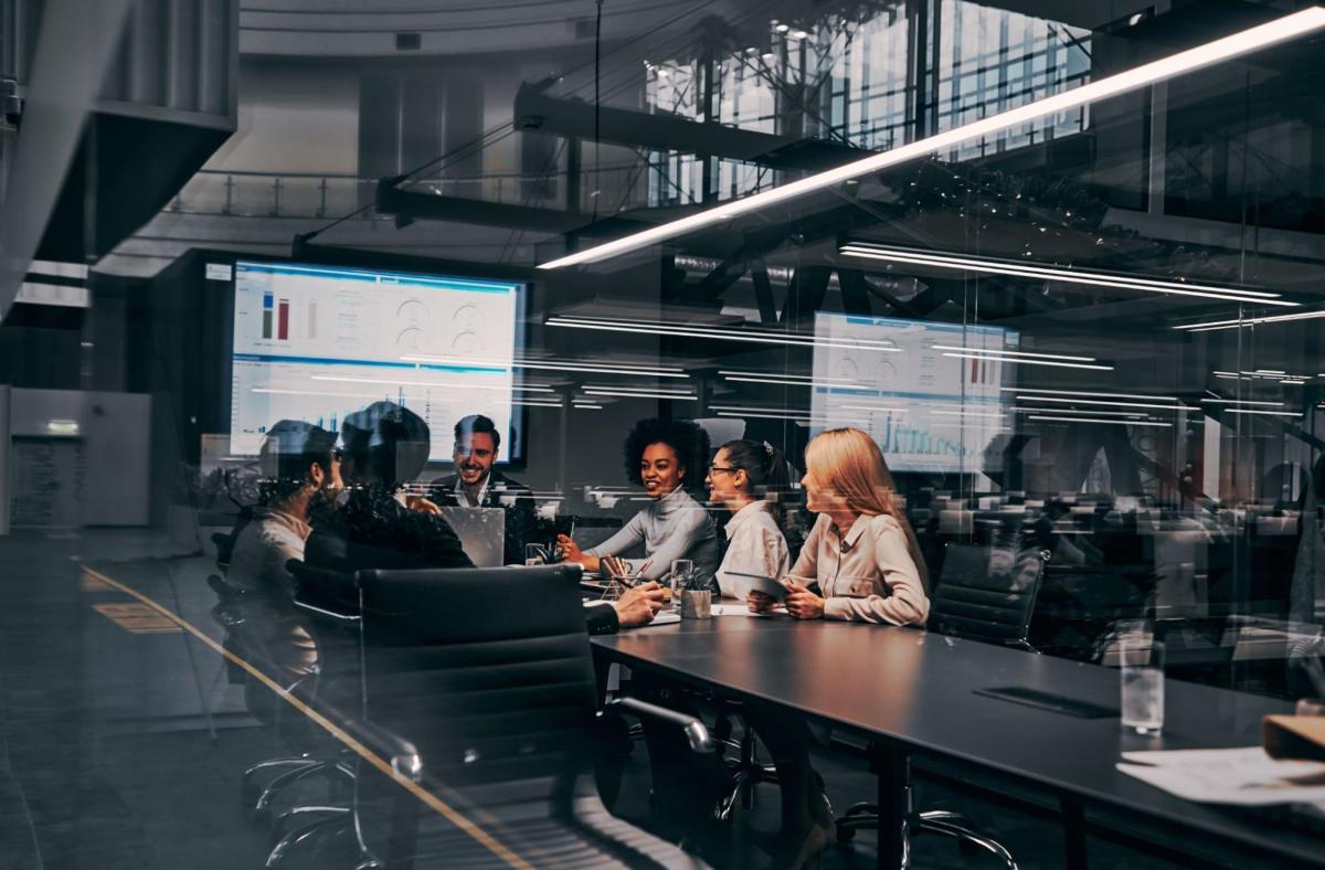 Photo of a group of co-workers seated around a conference room table with a big screen monitor in the back wall